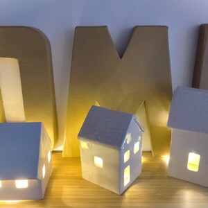 May include: Three white ceramic houses with light bulbs inside, sitting on a wooden surface in front of the letters "HOME" made of cardboard.