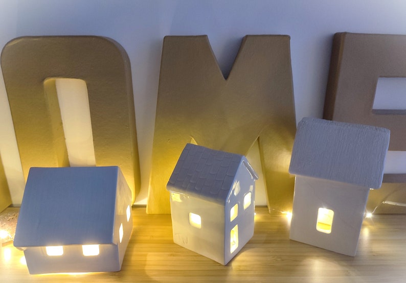 May include: Three white ceramic houses with light bulbs inside, sitting on a wooden surface in front of the letters "HOME" made of cardboard.