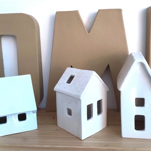 May include: Three white ceramic houses with cut-out windows, sitting in front of large brown cardboard letters that spell "HOME".