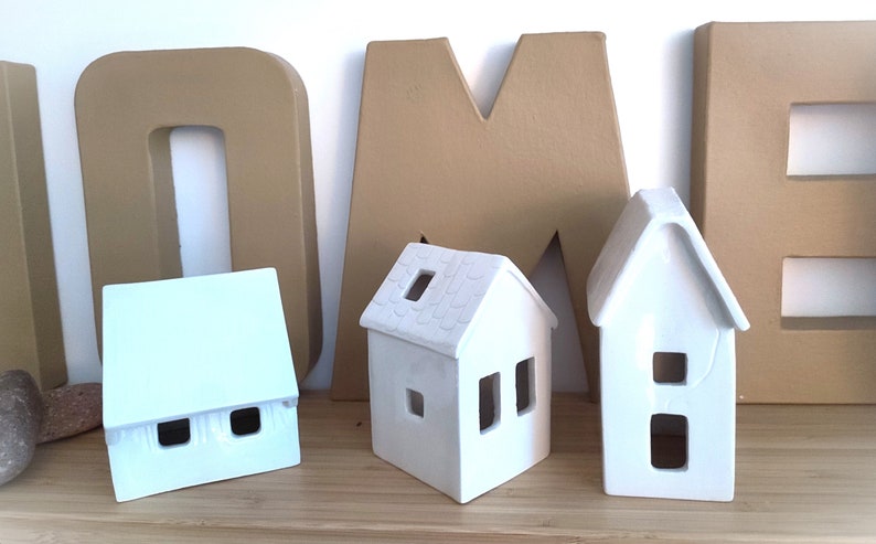 May include: Three white ceramic houses with cut-out windows, sitting in front of large brown cardboard letters that spell "HOME".