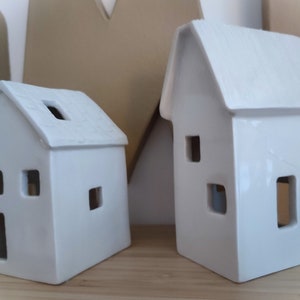May include: Two white ceramic houses with cut-out windows. The houses are different sizes and have a rustic, farmhouse style.