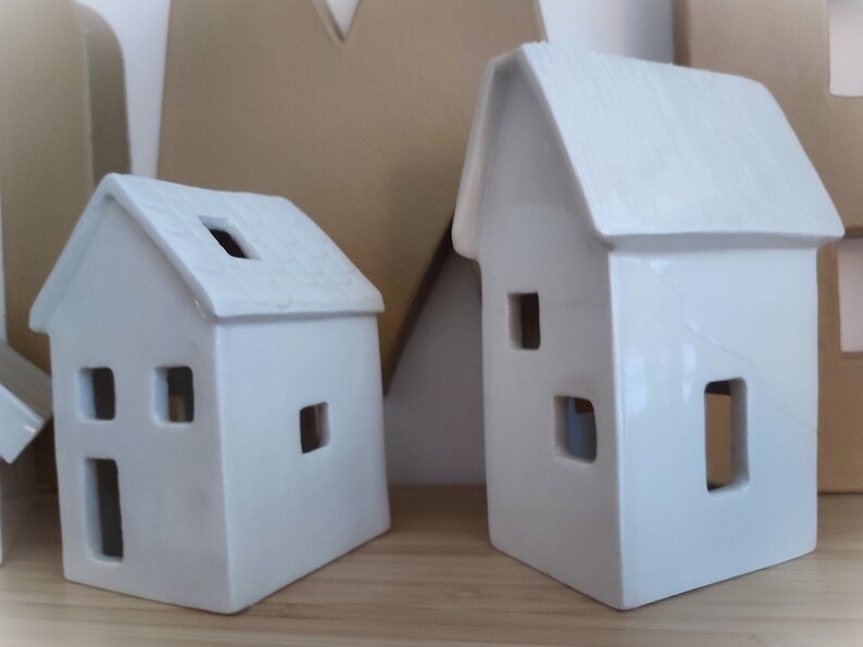May include: Two white ceramic houses with cut-out windows. The houses are different sizes and have a rustic, farmhouse style.
