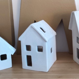 May include: Three white ceramic houses with cut-out windows and doors. The houses are different sizes and have a rustic, handmade look.