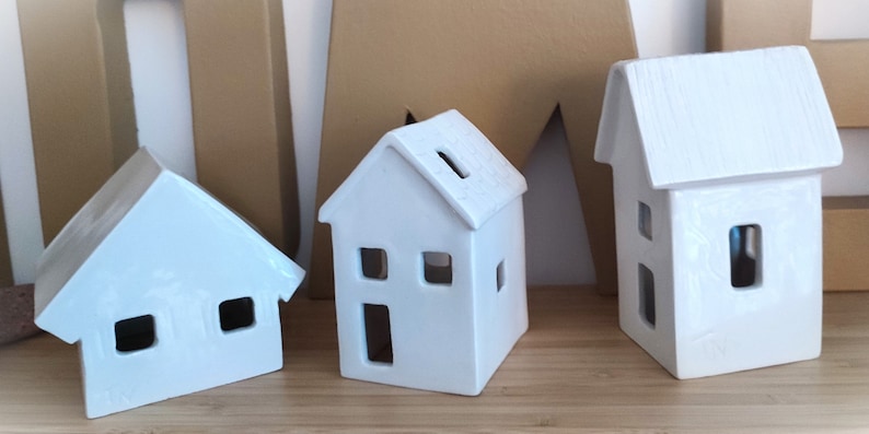 May include: Three white ceramic houses with cut-out windows and doors. The houses are different sizes and have a rustic, handmade look.
