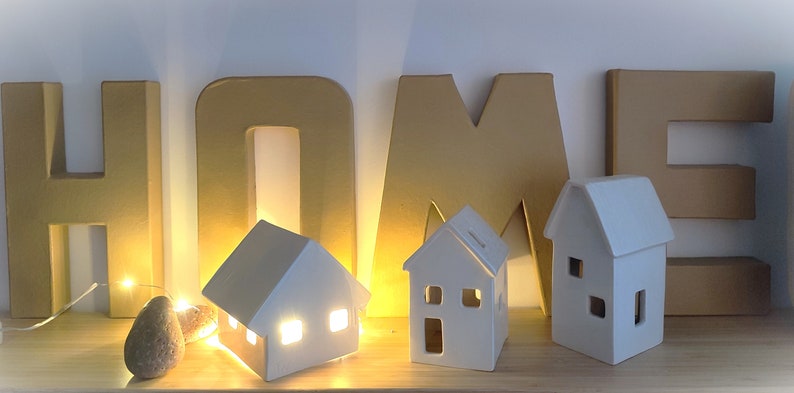 May include: White ceramic houses with lit windows, arranged in front of gold cardboard letters spelling "HOME".