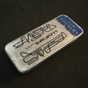 May include: A silver metal rectangular box with a black and white design and the text "...UNK 3117 FF." on the front. The top of the box has a blue label with the text "D71117 <" in white.