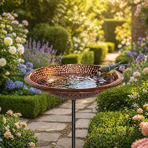 May include: A copper-colored bird bath with a hammered texture, filled with water. A small bird with blue, yellow, and white feathers is drinking from the water. The bird bath is on a black metal stand, surrounded by a lush garden.