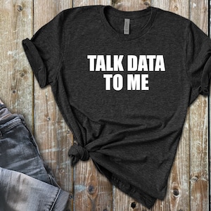 Op de afbeelding: Een donkergrijs T-shirt met de tekst "TALK DATA TO ME" in wit gedrukt op de voorkant.