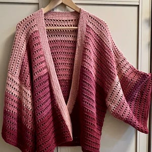 Puede incluir: Un cárdigan de ganchillo hecho a mano, abierto por delante, en tonos rosas y rojos. El cárdigan presenta un diseño de cuello en V y mangas anchas. El patrón de ganchillo crea una textura abierta y aireada. La prenda se exhibe en una percha de madera.