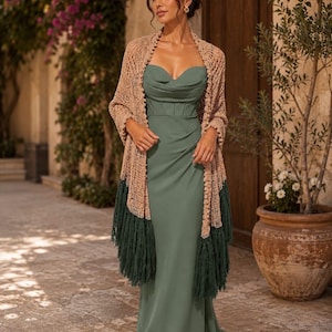 Può includere: Un abito lungo color verde salvia con scollo a barchetta e corpetto strutturato, abbinato a uno scialle beige a maglia aperta con motivo testurizzato e lunghe frange verde scuro che pendono lungo i lati del capo.
