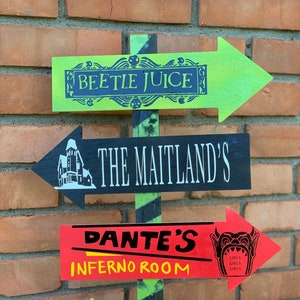 May include: A wooden directional sign with green, black, and red arrows. The arrows point in different directions and feature text and graphics from the movie Beetlejuice, including "Beetle Juice," "The Maitland's," "Dante's Inferno Room," and "Sandworms."