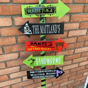 May include: A directional signpost with five arrow-shaped signs. The signs are painted in various colors, including green, black, and red, and feature text such as "BEETLE JUICE" and "SANDWORMS". The signpost is decorated with black and green stripes.
