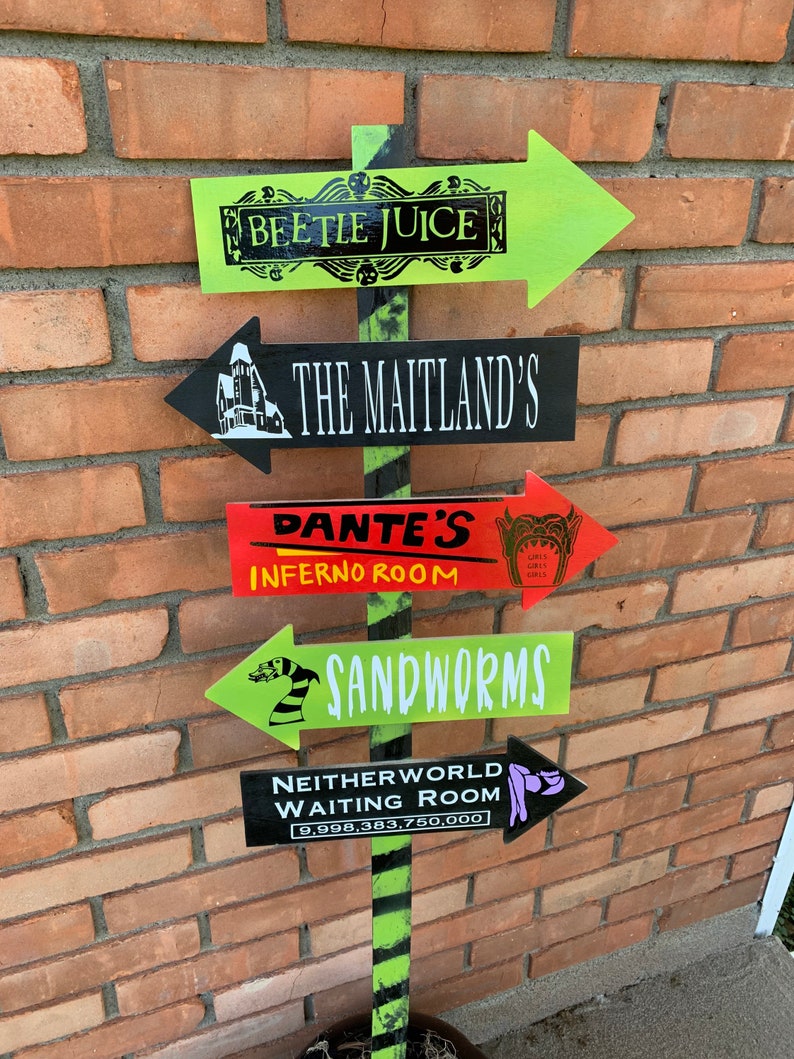 May include: A directional signpost with five arrow-shaped signs. The signs are painted in various colors, including green, black, and red, and feature text such as "BEETLE JUICE" and "SANDWORMS". The signpost is decorated with black and green stripes.