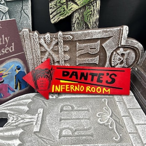 May include: A collection of Halloween-themed decorations, including a red sign that reads "Dante's Inferno Room" with a skull graphic, a book titled "Handbook for the Recently Deceased", and various tombstone-shaped ornaments.