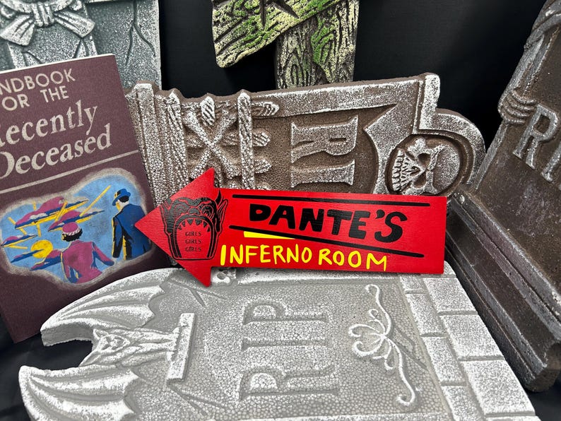 May include: A collection of Halloween-themed decorations, including a red sign that reads "Dante's Inferno Room" with a skull graphic, a book titled "Handbook for the Recently Deceased", and various tombstone-shaped ornaments.