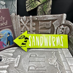 May include: A lime green arrow-shaped sign with the word "SANDWORMS" in white text, and a black and yellow sandworm illustration. The sign is placed on a gray tombstone, with a book and other tombstones in the background. Halloween decor.