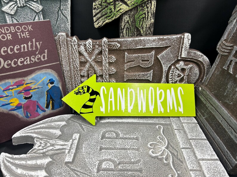 May include: A lime green arrow-shaped sign with the word "SANDWORMS" in white text, and a black and yellow sandworm illustration. The sign is placed on a gray tombstone, with a book and other tombstones in the background. Halloween decor.