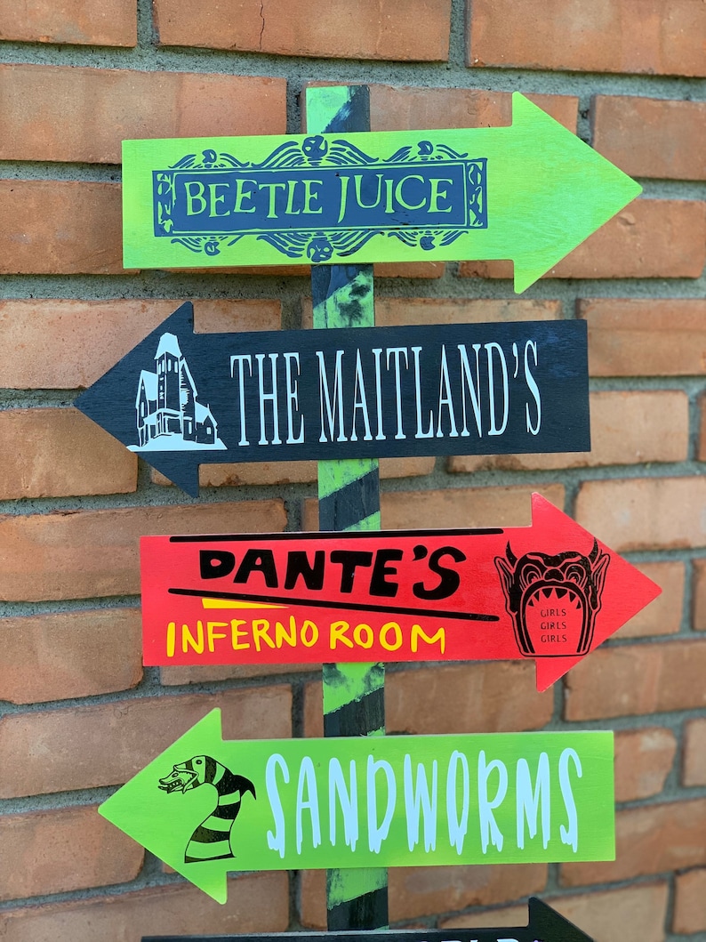 May include: A wooden directional sign with green, black, and red arrows. The arrows point in different directions and feature text and graphics from the movie Beetlejuice, including "Beetle Juice," "The Maitland's," "Dante's Inferno Room," and "Sandworms."