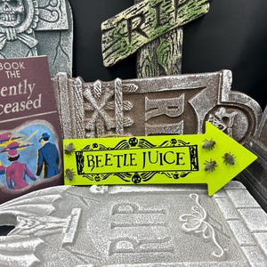 May include: A Halloween-themed display featuring a book titled "Handbook for the Recently Deceased" and a neon green arrow sign that reads "BEETLE JUICE". The sign has decorative skulls and insect accents. Tombstone props are in the background.
