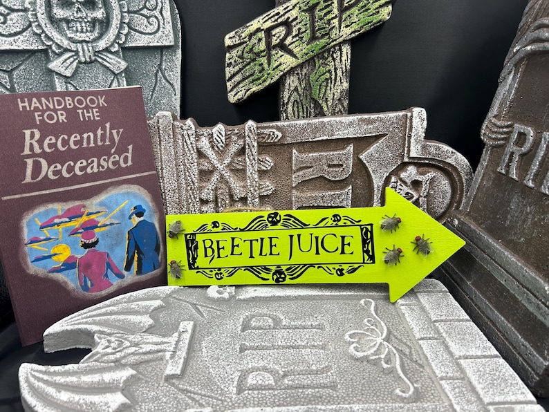 May include: A Halloween-themed display featuring a book titled "Handbook for the Recently Deceased" and a neon green arrow sign that reads "BEETLE JUICE". The sign has decorative skulls and insect accents. Tombstone props are in the background.
