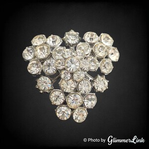 May include: A silver-toned brooch with a heart-shaped design. The brooch is covered in numerous clear, round, and faceted rhinestones of varying sizes, creating a sparkling effect. The rhinestones are set closely together, and the brooch is against a black background.