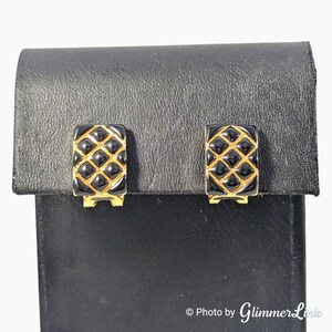 May include: A pair of black and gold clip-on earrings. The earrings have a rectangular shape with a quilted pattern in black enamel and gold-tone metal. The earrings are displayed on a black leatherette stand.