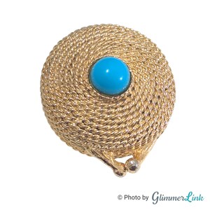 May include: A round gold-tone brooch featuring a textured, coiled rope design. The center of the piece is set with a smooth, polished, bright turquoise blue cabochon stone. The base of the circular frame includes a small, hinged metal clasp mechanism.