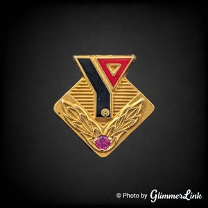 May include: A gold-toned pin featuring the YMCA logo. The pin has a diamond shape with a red and black triangle at the top, a black vertical line, and a small, round, pink gemstone at the bottom. The pin has a gold leaf design.