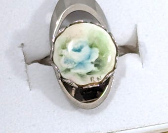 May include: A silver-toned ring featuring an oval-shaped setting with a hand-painted porcelain inlay. The inlay displays a soft blue rose surrounded by green foliage on a cream background, with the initials RN clearly visible in the lower right corner.
