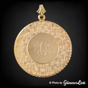 May include: A gold-colored pendant with a textured, circular design. The pendant features a smaller, smooth circle in the center with an engraved monogram. A decorative bail is at the top for hanging. The overall design is vintage and elegant.