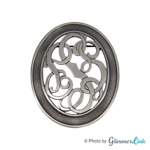 May include: Silver oval brooch with a cutout design of intertwined letters. The brooch has a textured border.