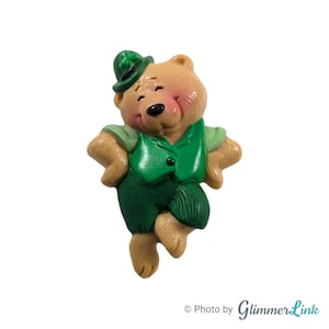 May include: A green and brown figurine of a bear wearing a green hat and a green vest. The bear is standing on one leg and has its other leg raised.