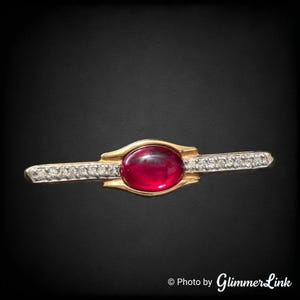 Peut inclure: Une broche barre horizontale en or, ornée d'une pierre précieuse centrale ovale rouge foncé taillée en cabochon. Les bras allongés de la broche sont agrémentés d'une rangée de petites pierres claires et scintillantes sur le cadre métallique.