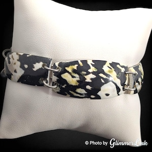 May include: A bracelet with a black, white, and yellow speckled pattern. The bracelet is made of several rectangular segments connected by silver-colored metal links. The segments have a glossy finish and are resting on a white surface.
