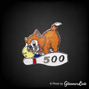 May include: A silver pin with an orange cat holding a yellow bowling ball and a white bowling pin with the number 500 on it.
