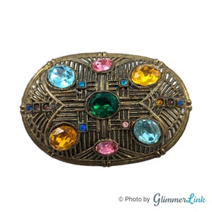 May include: An oval-shaped brooch with a gold-toned metal frame and a geometric design. The brooch is adorned with colorful rhinestones in shades of blue, green, pink, and yellow.