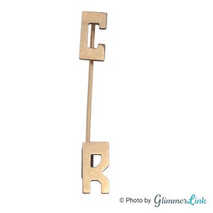May include: A gold-colored pin featuring the letters "C" and "R" connected by a thin, straight rod. The letters are block-style, with the "C" at the top and the "R" at the bottom. The pin is set against a light background.