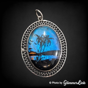 May include: An oval silver pendant with a delicate filigree design. The pendant features a blue and black butterfly wing with a silhouette of palm trees against a sunset.