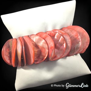 May include: A coral-colored bracelet composed of large, flat, round beads. The beads have a pearlescent sheen and are strung together to form a flexible band. The bracelet is displayed on a white, square pillow against a black background.