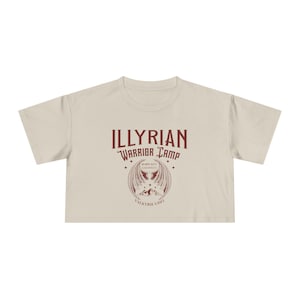Puede incluir: Top corto color crema con el texto "ILLYRIAN WARRIOR CAMP" en granate. Debajo, un diseño alado con las palabras "VALKYRIE UNIT". La camiseta tiene mangas cortas y cuello redondo.