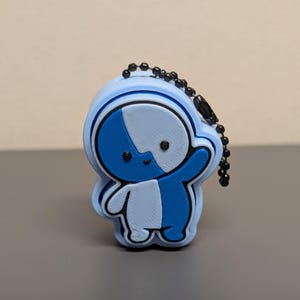 May include: A small, light blue and dark blue character-shaped keychain with a split-color design. The figure features a rounded head, a small body, and a black beaded chain attached to the top. The surface has a layered, textured appearance.