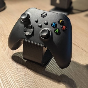 May include: A black Xbox controller with a black stand. The controller has a green, blue, yellow, and red button.