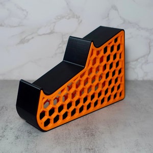 May include: A black and orange 3D printed stand with a honeycomb design. The stand has two levels and is designed to hold items like phones or controllers.
