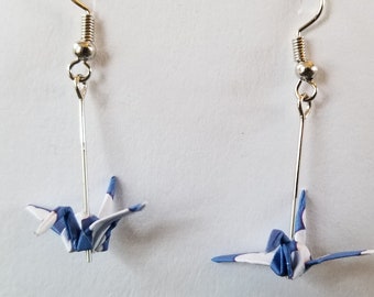 May include: A pair of origami crane earrings. Each earring features a small, folded paper crane in blue and white, suspended from a gold-colored hook. The cranes are detailed with visible folds, creating a delicate and artistic look.