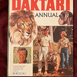 May include: A vintage "Daktari Annual" book cover. The cover features a lion, a chimpanzee, and a man and woman with a tiger. The title "DAKTARI ANNUAL" is in large red letters. The book is an authorised edition based on the MGM television film series.