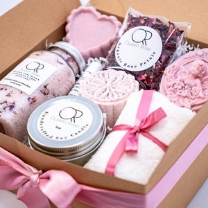 May include: A brown gift box with a pink ribbon tied around it. The box is filled with a variety of bath and body products, including bath salts, a candle, soap, and rose petals. The products are all pink and white and have a floral theme.
