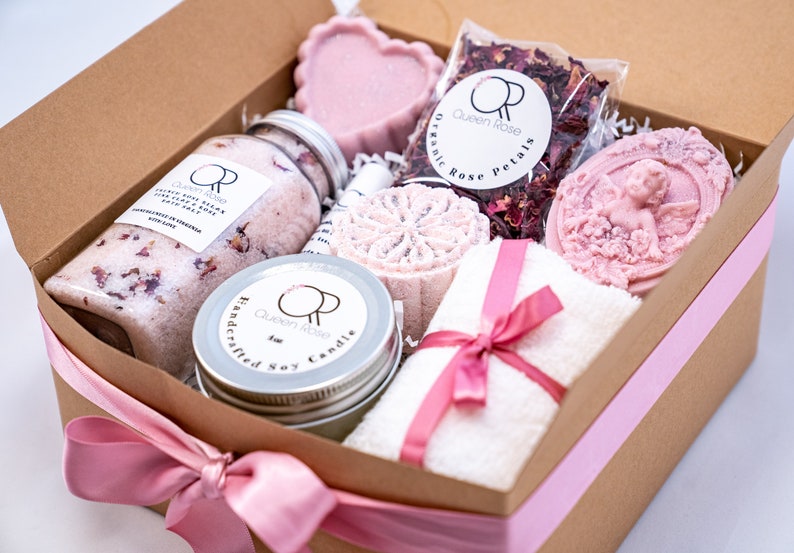 May include: A brown gift box with a pink ribbon tied around it. The box is filled with a variety of bath and body products, including bath salts, a candle, soap, and rose petals. The products are all pink and white and have a floral theme.