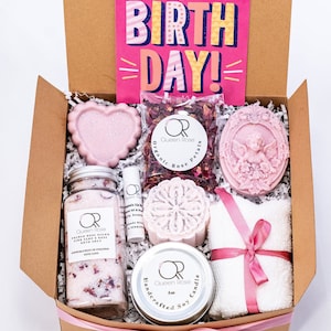 May include: A brown gift box tied with a pink ribbon, filled with bath and body products. The box contains bath salts, a candle, a bath bomb, a bar of soap, a card that says "Happy Birthday!" and a bag of rose petals.