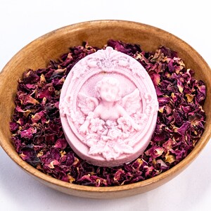 May include: A pink oval-shaped soap with a cherub design in a wooden bowl filled with dried rose petals.