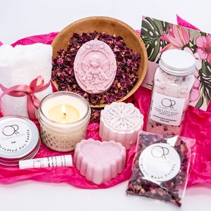May include: A pink and white spa gift set with a candle, bath bombs, bath salts, rose petals, and a hand towel. The set is packaged in pink tissue paper and includes a card with a floral design.